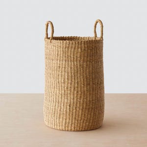 May include: A large, natural woven basket with two handles. The basket is made of a light brown material and has a cylindrical shape.