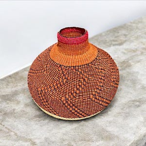 May include: A woven basket with a round, bulbous shape. The basket is made of natural materials and has a brown and black pattern. The top of the basket is trimmed with a pink band.
