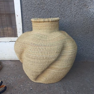 Peut inclure: Un grand panier rond et bulbeux, tissé à la main, de couleur naturelle. Le panier est fait de fibres naturelles tissées serrées et a une surface légèrement texturée.