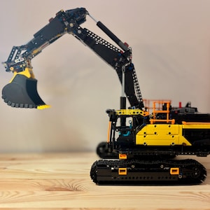 Peut inclure: Une maquette d'excavatrice noire et jaune détaillée avec un grand godet. L'excavatrice a un long bras et est posée sur une base à chenilles. Le modèle est sur une surface en bois.