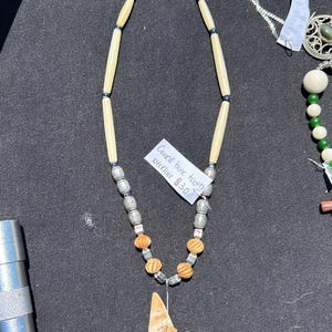 May include: A handcrafted necklace featuring bone-shaped beads, silver-toned metal beads, and orange wooden beads. The pendant is a carved bone tooth with a landscape design. The tag reads "Carved bone tooth Necklace".