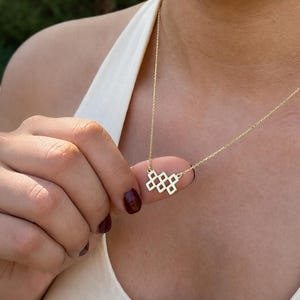 May include: A gold-toned necklace featuring a delicate chain and a pendant composed of four interconnected diamond-shaped geometric cutouts arranged in a horizontal, slightly staggered pattern, resting against a white ribbed tank top.