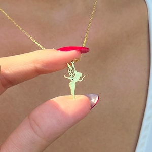 May include: A gold-colored necklace featuring a delicate fairy pendant. The fairy has detailed wings and is suspended from a fine chain. The necklace is being held up by a person's fingers, showcasing the pendant's design.