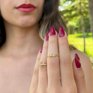 May include: Two gold-colored rings, one spelling "Chloe" and the other "Sophie", are displayed on a person's fingers. The rings are in a delicate script font. The person's nails are painted a deep red color.
