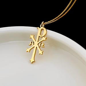 May include: A gold Chi Rho pendant necklace, a Christian symbol formed from the first two letters of the Greek word "Christos." The pendant is suspended from a fine gold chain. The background is white.