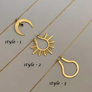 May include: Three gold necklaces with different pendant styles. The first necklace has a crescent moon pendant with a small blue gemstone. The second necklace has a sunburst pendant. The third necklace has a teardrop-shaped pendant.