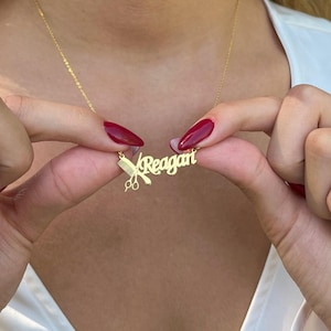 May include: A gold-colored necklace with the name "Reagan" in a cursive font. The pendant includes a comb and scissors, suggesting a hairstylist theme. The necklace is held by a person with red nail polish.