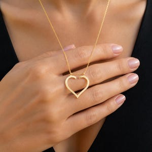 May include: A gold-toned necklace featuring a delicate chain and a pendant shaped like an open heart outline. The heart pendant is suspended from the chain at two points, creating a minimalist and clean aesthetic with a polished metallic finish.