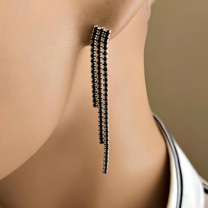 Op de afbeelding: Een goudkleurige oorbel met meerdere strengen kleine, zwarte edelstenen die naar beneden vallen. De oorbel is aan het oor bevestigd, met de strengen vrij hangend. Het ontwerp is elegant en geschikt voor formele gelegenheden.
