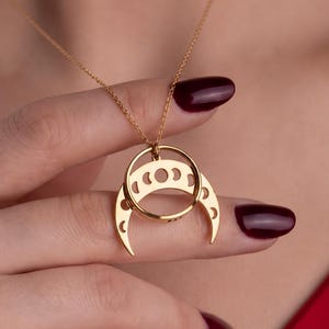 May include: A gold-toned necklace featuring a pendant with a circular ring frame and a crescent moon shape suspended below it. The crescent moon component displays a series of cut-out moon phase symbols, including full, half, and crescent shapes.