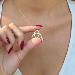 May include: A gold-colored pendant necklace featuring a Celtic knot design. The pendant is held between fingers with red nail polish. The necklace chain is delicate and also gold-colored. The background is a white top.