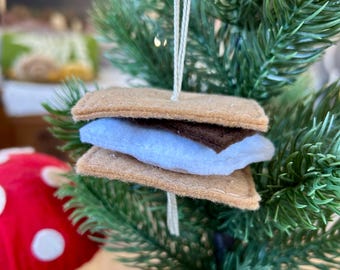 May include: A felt s'more ornament with a brown graham cracker, a white marshmallow, and a brown chocolate layer. The ornament is hanging on a green Christmas tree.