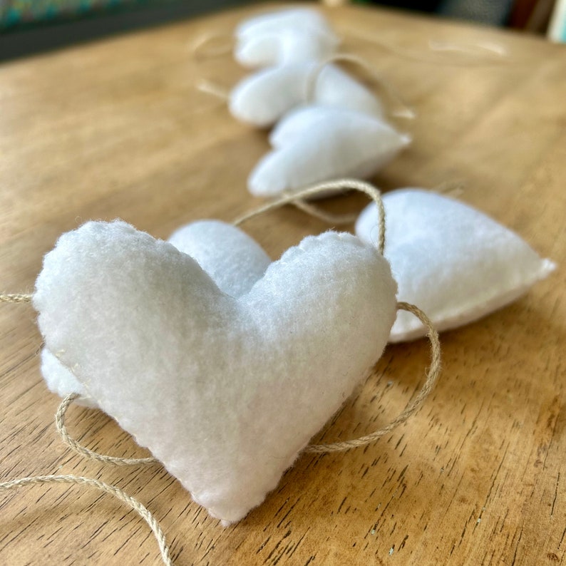 A garland of white felt hearts strung together with natural cord. The hearts are arranged in a row and are all the same size and shape. The hearts are soft and fluffy and have a slightly textured surface.