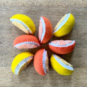 May include: A collection of eight felt fruit slices shaped like citrus wedges. The pieces are colored in bright yellow and vibrant orange, each featuring a white felt layer between the peel and the segmented fruit interior, arranged in a circular pattern.