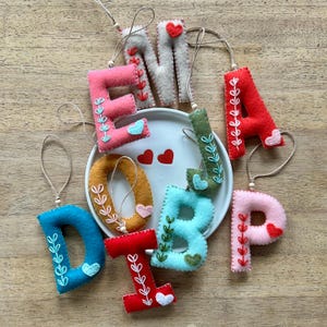 May include: Felt letter ornaments in various colors with embroidered hearts and leaves. The letters spell out "DIPJBE".