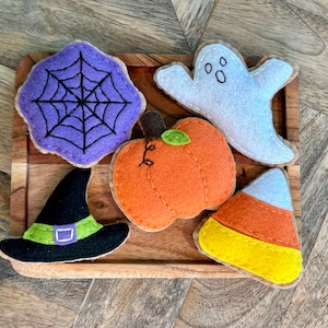 May include: Five felt Halloween-themed ornaments: a purple spiderweb, a white ghost with two black eyes, an orange pumpkin with a green stem, a black witch's hat with a green band, and a yellow and orange candy corn.