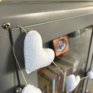 A string of white felt hearts hanging from a gray metal surface. The hearts are connected by a thin, natural twine string.