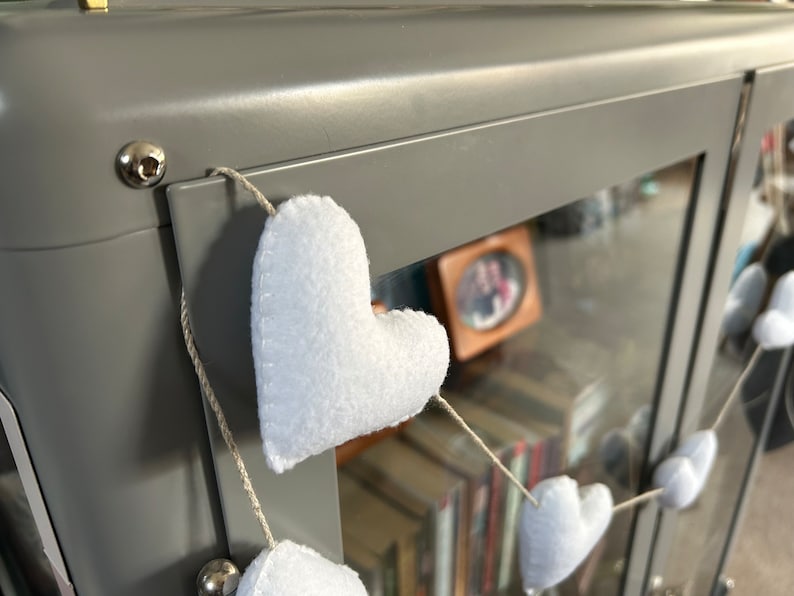 A string of white felt hearts hanging from a gray metal surface. The hearts are connected by a thin, natural twine string.