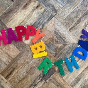 May include: A colorful felt banner spelling "HAPPY BIRTHDAY" in rainbow hues. The letters are strung on a white cord, perfect for birthday celebrations. The banner is displayed on a wooden surface.