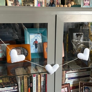 A white felt heart garland hanging in front of a glass cabinet with books, pictures, and other items inside. The garland is strung with natural cord and has a white felt heart at each end. The garland is hanging in front of a gray cabinet with glass doors.
