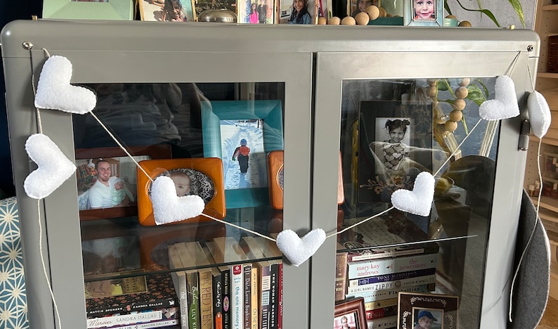 A white felt heart garland hanging in front of a glass cabinet with books, pictures, and other items inside. The garland is strung with natural cord and has a white felt heart at each end. The garland is hanging in front of a gray cabinet with glass doors.