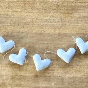 A string of white felt hearts connected by natural cord. The hearts are arranged in a curved line on a wood surface.