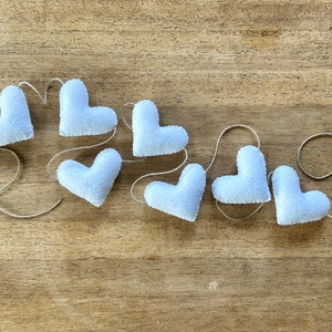 A string of white felt hearts connected with natural cord. The hearts are arranged in a curved line, with the string running through the top of each heart.