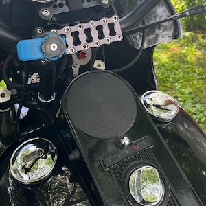 May include: Close-up of a black motorcycle with chrome accents. The handlebars feature a blue phone mount and a silver chain-like accessory. The fuel tank has a black pad and chrome fuel caps. The motorcycle is parked outdoors.