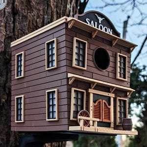 Könnte beinhalten: Ein braunes, zweistöckiges Vogelhaus, das einem Saloon nachempfunden ist. Es hat ein Schild mit der Aufschrift "SALOON" und verfügt über mehrere Fenster, ein rundes Eingangsloch und dekorative Elemente wie ein Holzrad und ein Fass. Das Vogelhaus ist an einem Baum befestigt.