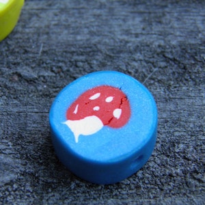 May include: A blue and white clay bead with a red mushroom design. The bead is round and has a hole in the center.
