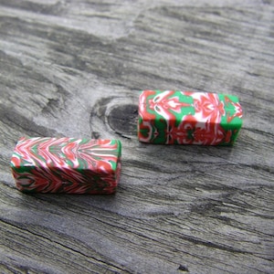 May include: Two small rectangular polymer clay beads with a red, white, and green marbled pattern. The beads are on a wooden surface.