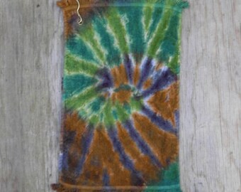 May include: A rectangular hand towel featuring a tie-dye spiral pattern in shades of green, brown, and purple. The towel has fringed edges on the top and bottom and includes a small metal hook attached to the top left corner for hanging purposes.