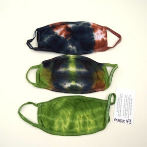 Op de afbeelding: Drie tie-dye gezichtsmaskers in verschillende kleuren: blauw, groen en bruin. De maskers zijn handgemaakt en wasbaar. De tekst op de kaart luidt: "MADE BY HIPPIES Tie Dyes are hand dyed using Procton Dye so they are color fast and can be washed with your regular laundry. To get 20% off your next Zay purchase enter coupon code TheRippyDiscount MASK 43"
