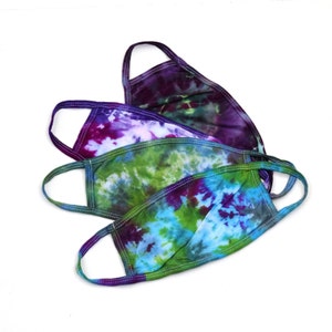 May include: Three tie-dye face masks in shades of purple, green, and blue. The masks are made of cotton and have elastic ear loops.