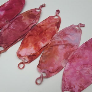 Op de afbeelding: Vijf tie-dye gezichtsmaskers in tinten van roze, rood en oranje. De maskers zijn gemaakt van een zachte, ademende stof en hebben verstelbare oorlussen.