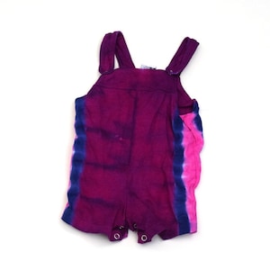 Puede incluir: Un pelele de bebé teñido anudado en tonos morado, azul y rosa. El pelele tiene tirantes ajustables y cierres de botón a presión en la parte inferior. La prenda está hecha de tela suave y está diseñada para la comodidad y el estilo.