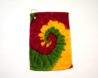 May include: A rectangular towel featuring a spiral tie-dye pattern in shades of deep red, bright yellow, and forest green. The towel includes a metal grommet in the top left corner with a gold-toned metal ring attached for hanging purposes.