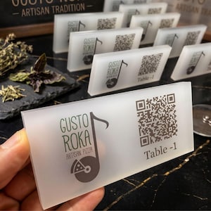 May include: A rectangular, frosted acrylic table sign featuring the black printed logo for Gusto Roka Artisan Pizza, a musical note icon, a QR code, and the text Table-1. The sign is held in the foreground with several identical signs visible behind.