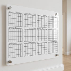 May include: A white acrylic wall calendar with a grid layout for each month of the year. The calendar is held in place by four silver-coloured mounting hardware pieces. Each month's grid is labelled with the month's name.