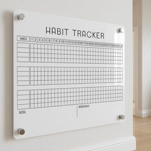 以下が含まれることがあります： 「HABIT TRACKER」の文字が上部に書かれた白いアクリル製の習慣トラッカー。習慣を追跡するためのグリッドがあり、月の各日は上部に番号が振られています。「NEVER GIVE UP」の文字が下部にあります。