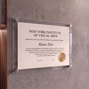Op de afbeelding: Een transparante, aan de muur gemonteerde documentenlijst van acryl met een certificaat van het New York Institute of Visual Arts voor een Bachelor of Fine Arts in Visual Communication Design voor Klara Tarr, met vier zilveren metalen houders.