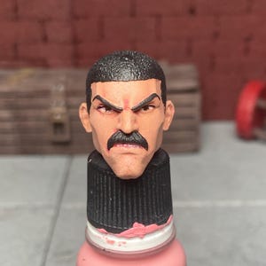May include: A detailed, painted action figure head with a stern expression, dark hair, and a prominent mustache. The head is mounted on a pink paint bottle with a black cap. The background features a brick wall and a wooden crate.