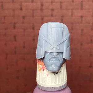 May include: A light blue, unpainted miniature head with a helmet, resting on a purple bottle. The head has a stern expression and is likely a collectible or model kit piece. The background is a red brick wall.