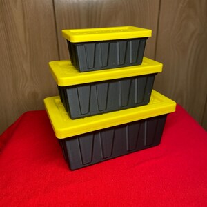 May include: Three black storage containers with yellow lids are stacked on a red surface. The containers have a rectangular shape with vertical grooves on the sides. The lids are a bright yellow color.