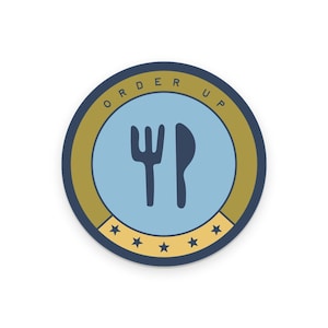 May include: Round sticker with a blue centre showing a fork and knife. The sticker has a gold border with the words "ORDER UP" in a circular pattern. Small gold stars are at the bottom of the sticker.