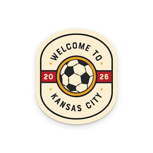 May include: Cream-colored oval sticker with a soccer ball graphic. The sticker features the text "WELCOME TO KANSAS CITY" in a circular design, with the numbers "20" and "26" on red bars. A yellow circle surrounds the soccer ball.