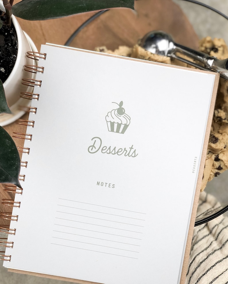 以下が含まれることがあります： 緑色のカップケーキのイラストと「Desserts」という文字が書かれた白いスパイラルバインダーノート。ノートには罫線が入ったページがあり、メモを書くことができます。