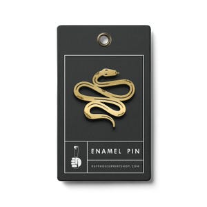 May include: A gold-coloured enamel pin shaped like a snake, presented on a black tag with a white border. The tag features the text "ENAMEL PIN" and the website address "RUFFHOUSEPRINTSHOP.COM". This pin is a decorative accessory.
