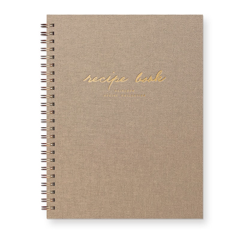 Spiral bound keepsake recipe book with tan linen cover. Gold foil font reads "recipe book heirloom recipe collection."