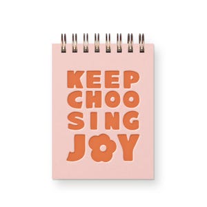 May include: A light pink spiral-bound notebook featuring the orange text "KEEP CHOOSING JOY" on the front cover. The letter "O" in the word "JOY" is replaced by a simple five-petaled flower shape. The notebook has a metallic wire binding at the top.